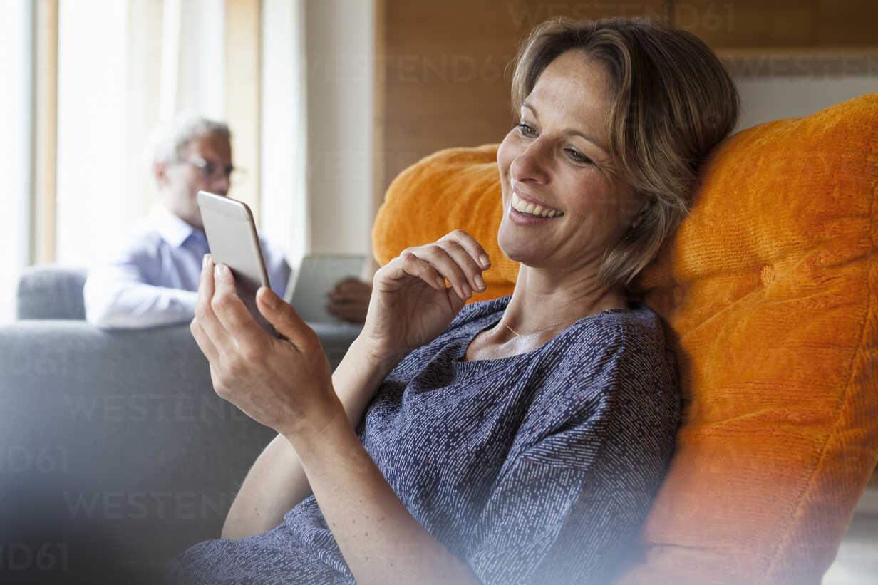 Smiling Woman At Home Looking At Cell Phone With Husband In Background Stockphoto