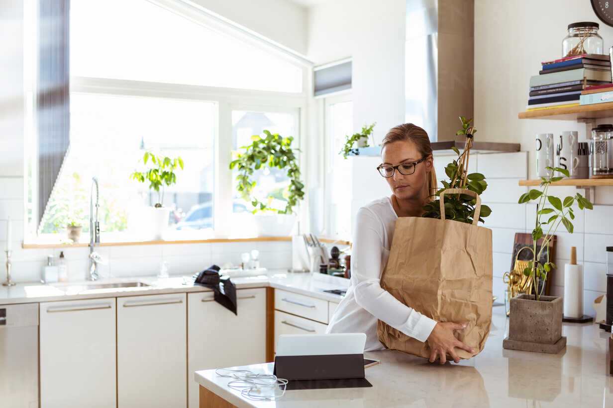Woman Holding Paper Bag While Looking At Digital Tablet In Kitchen