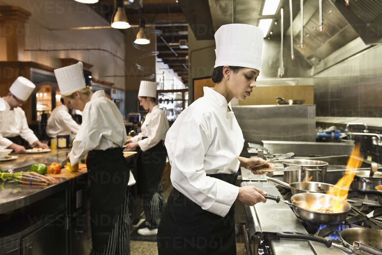 A Crew Of Chefs Working In A Commercial Kitchen With A Black Chef In The Foreground Sauteing Vegetables MINF09217 