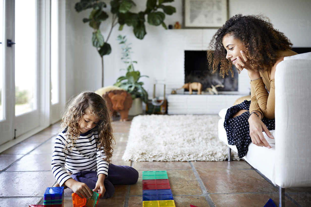 Mother Looking At Daughter Playing With Toy Blocks In Living Room Stockphoto