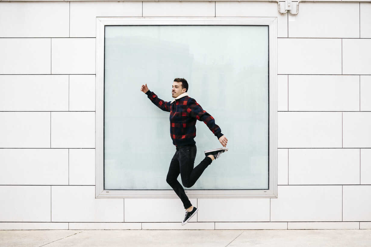 Young Man With Casual Clothes Jumping With A White Wall And A Large Window In The