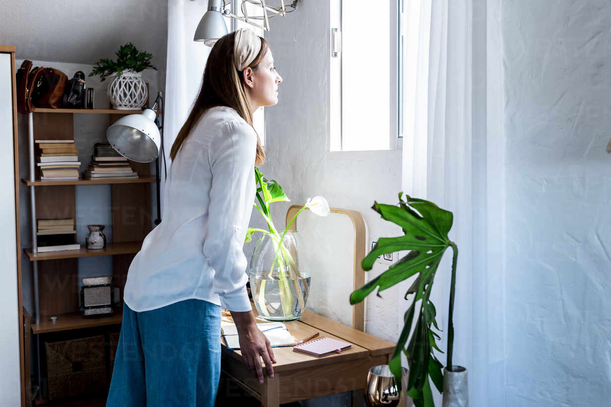 Woman Looking Through Window While Standing At Desk In Bedroom Stockphoto