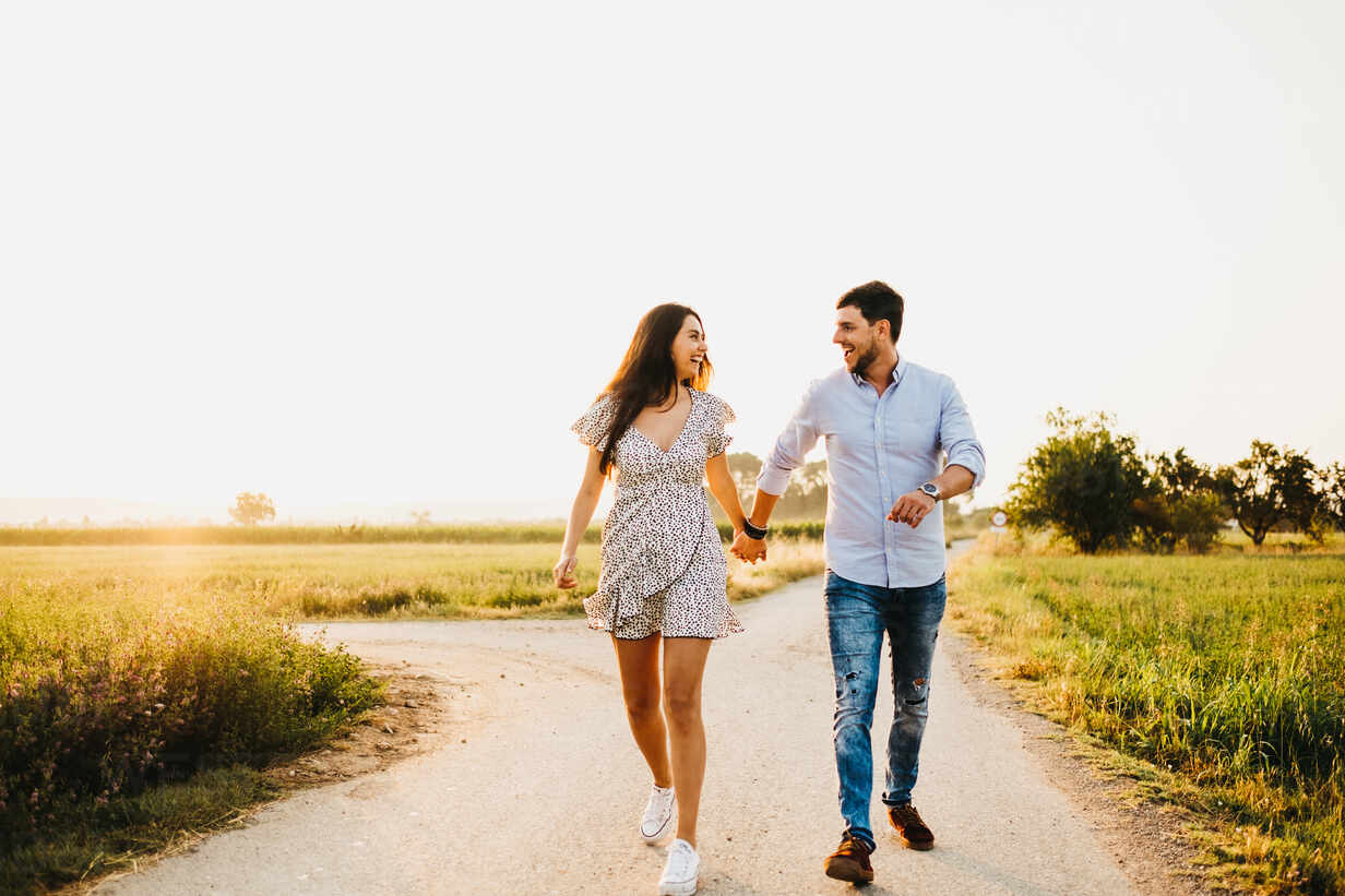 Cheerful Young Man And Woman Holding Hands And Walking On Field In Sunlight Stockphoto 